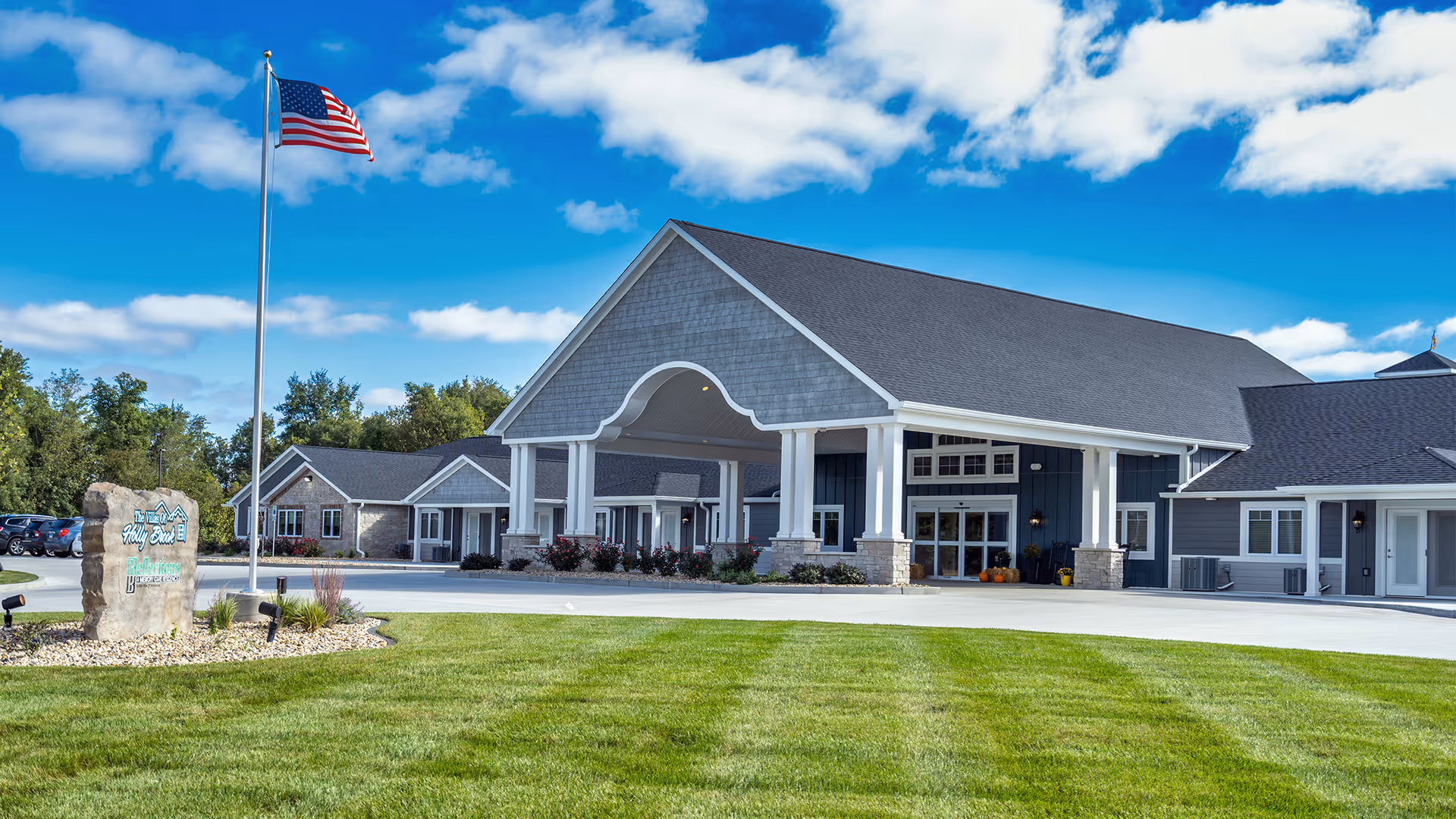 Front exterior view of Villas of Holly Brook Assisted Living & Memory Care building with a large covered entrance, an American flag on a flagpole, a stone sign with the facility name, and a well-maintained lawn under a partly cloudy blue sky.