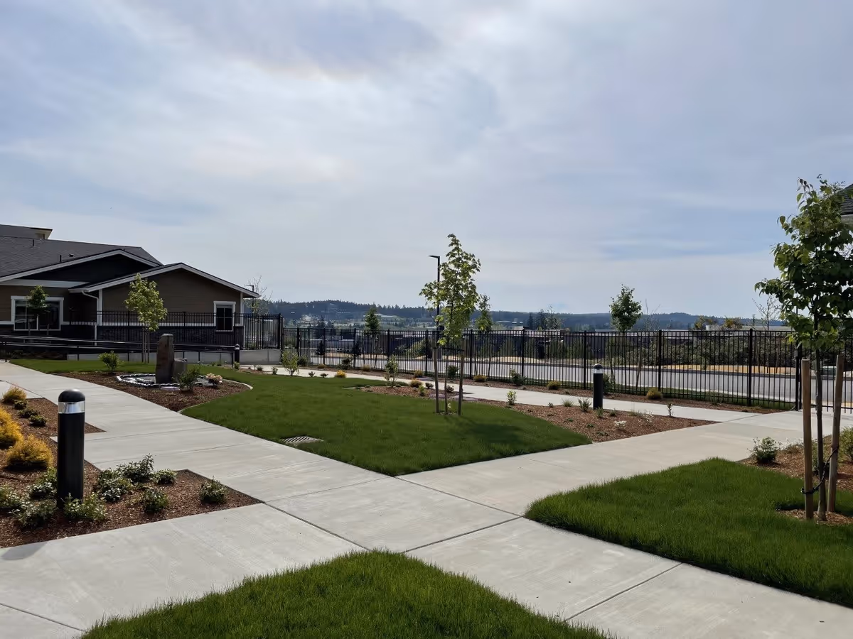 Outdoor garden area at Fieldstone Memory Care Silverdale with concrete walkways intersecting through well-maintained green grass, young trees, and landscaped shrubbery. A building is visible on the left side, and a black metal fence encloses the area with a view of distant hills under a partly cloudy sky.