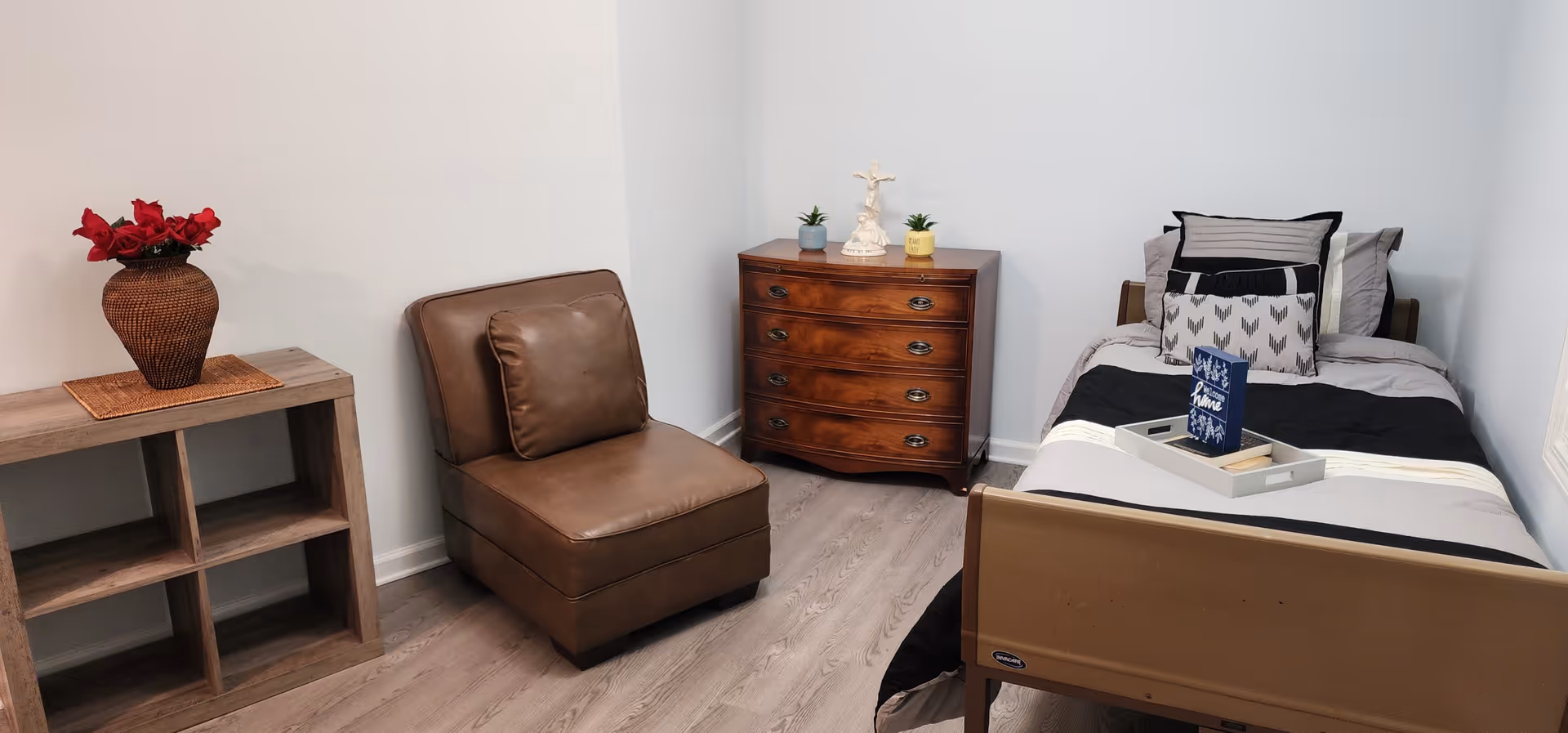 A cozy senior care bedroom with a single bed neatly made with pillows and a tray on top. There is a wooden chest of drawers with decorative items, a brown leather chair, and a small wooden shelf with a vase of red flowers. The room has light-colored walls and wood flooring.