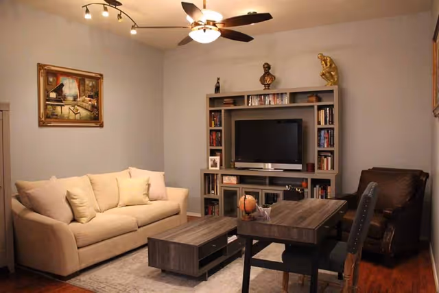 Cozy living room with a beige sofa, TV in a shelving unit, coffee table, dining table and armchair under a ceiling fan.