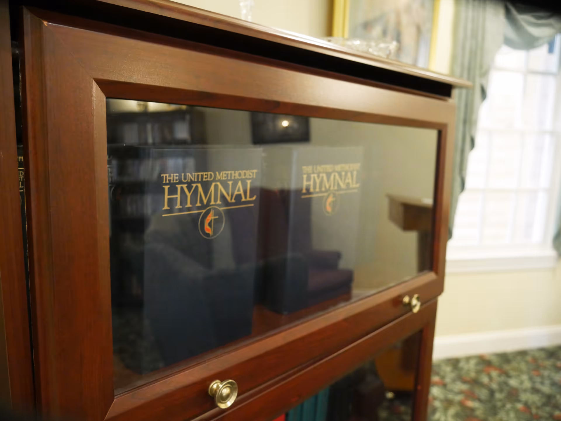 Close-up of a wooden glass-front cabinet displaying copies of "The United Methodist Hymnal" in a well-lit room.