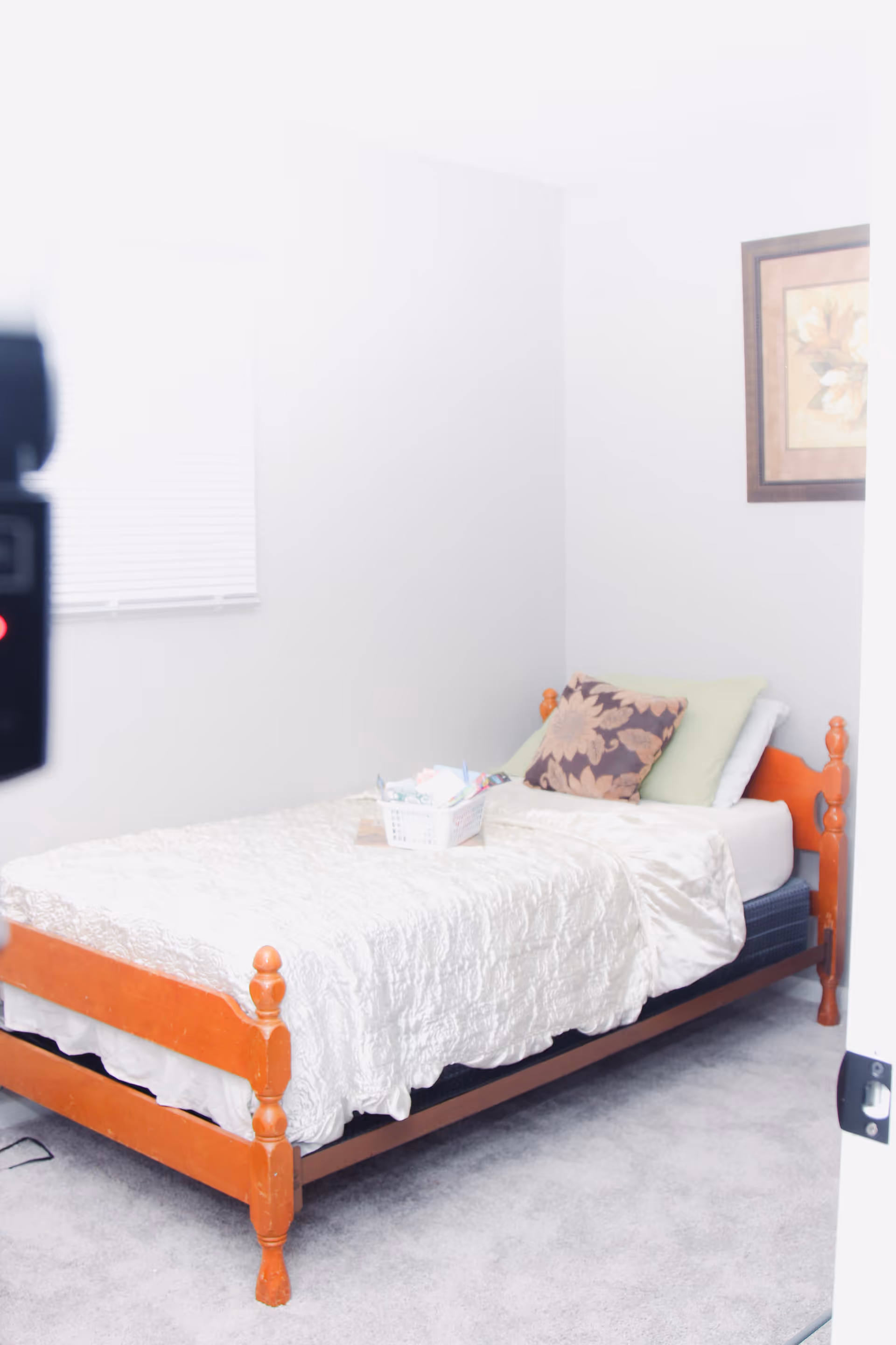 Small bedroom with a single wooden bed covered by a white quilt, pillows, a basket on the bed, a window with blinds, and a framed picture on the wall.