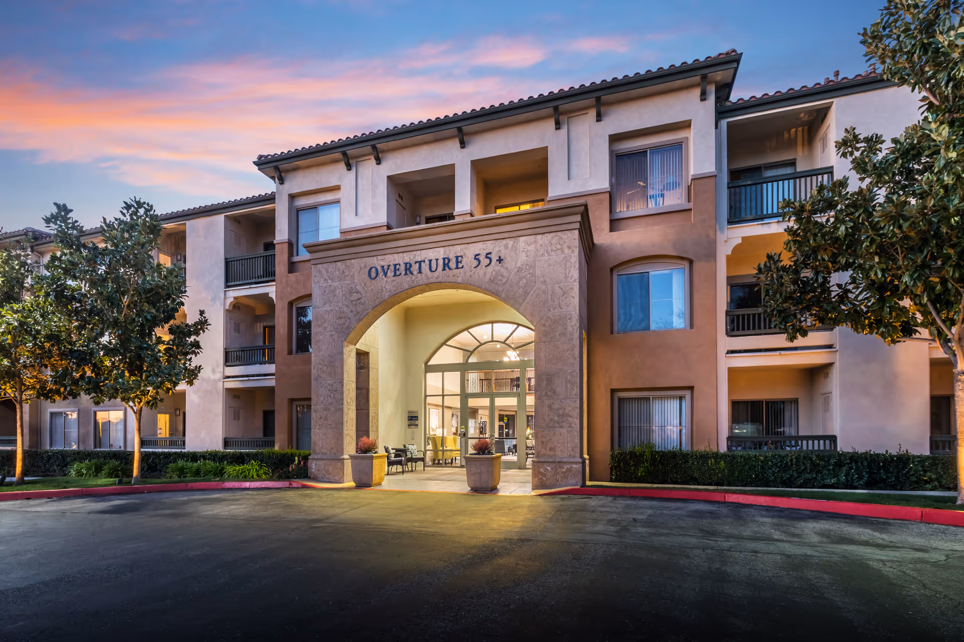 Exterior view of Overture Rancho Santa Margarita senior living facility at dusk, showing a three-story building with balconies, an arched entrance with the sign 'OVERTURE 55+', trees, and a paved driveway.