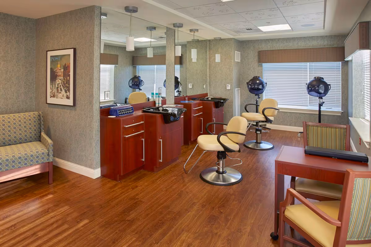 A senior living facility salon area with two beige salon chairs in front of wooden counters with sinks and mirrors. There are two blue hair dryers on stands near the windows with blinds. The room has wood flooring, a patterned couch, a framed picture on the wall, and a small table with two chairs.