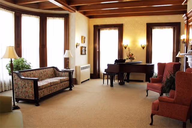 Sunlit sitting room with a floral sofa, upholstered armchairs, lamps, and a grand piano beside tall windows.