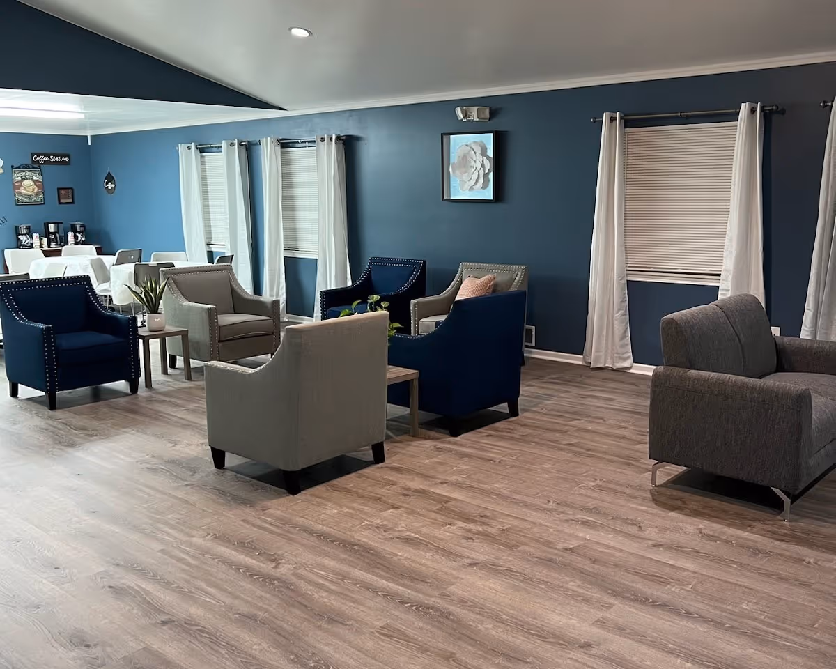 Spacious common lounge with multiple upholstered armchairs and small tables on wood-look flooring and teal walls.