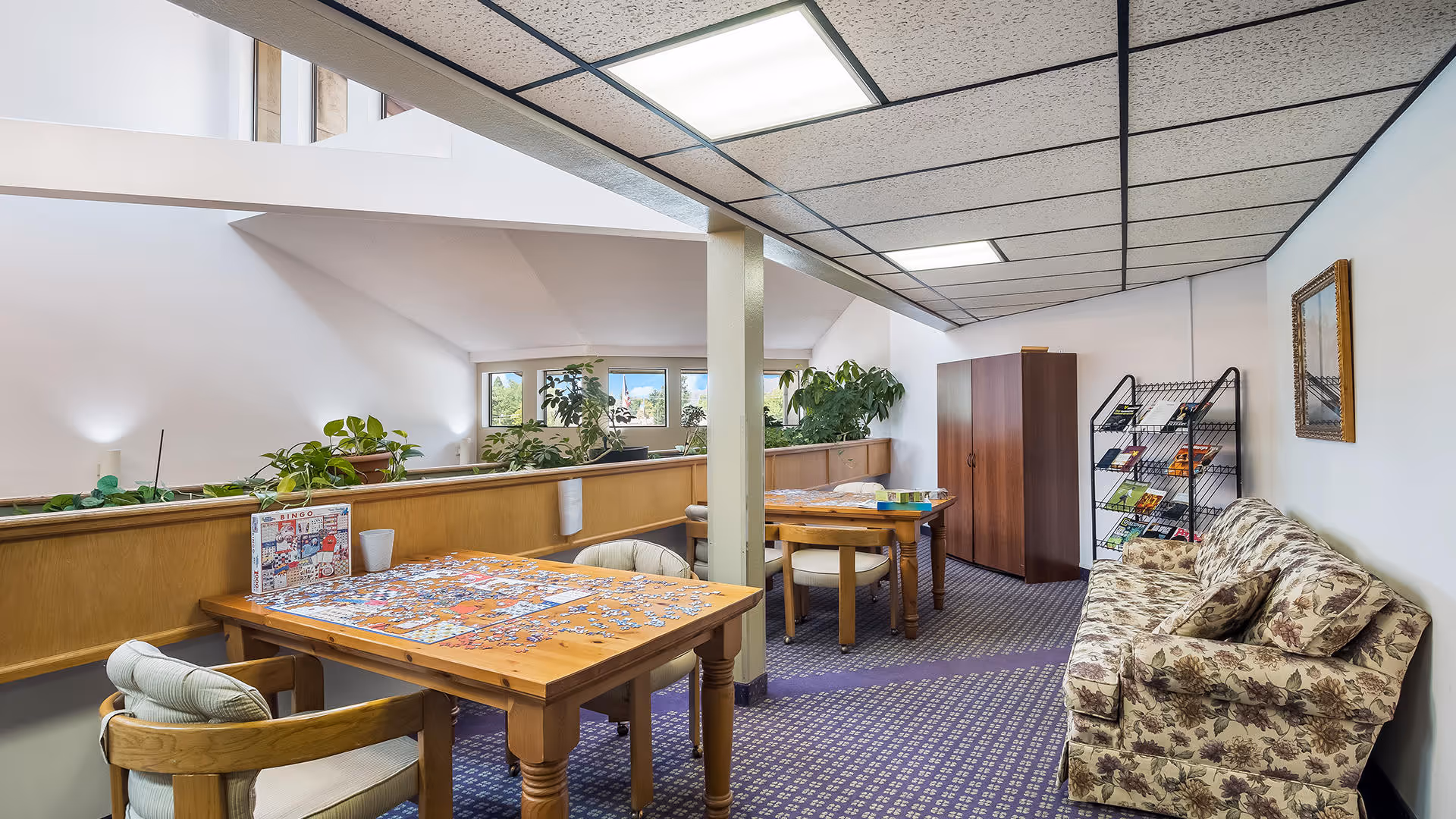A bright communal activity room with wooden tables covered in a jigsaw puzzle, a floral couch, plants, and shelving.