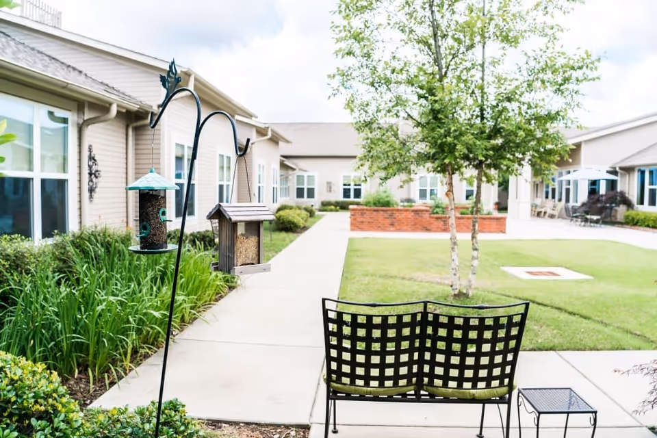 Outdoor courtyard area at Pinnacle Place Memory Care featuring a paved walkway, green grass, a small tree, a black metal bench with green cushions, a small side table, and two bird feeders hanging from a metal pole. The courtyard is surrounded by single-story beige buildings with multiple windows.