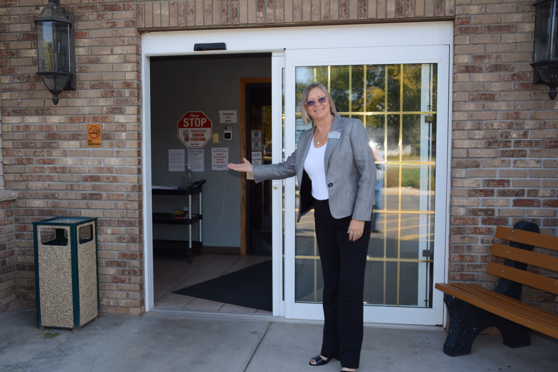 A woman stands at the sliding glass entrance of a brick building, smiling and gesturing toward the doorway.