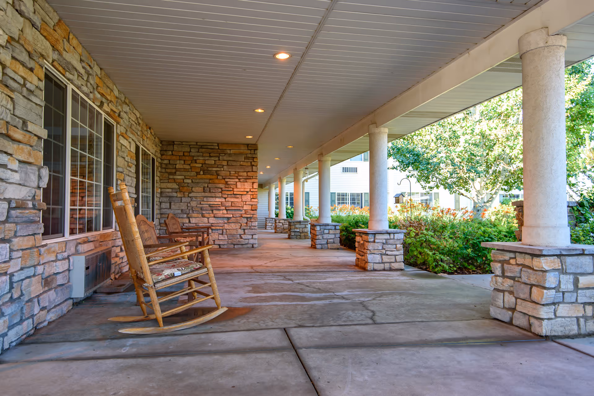 Covered outdoor porch area with stone pillars and walls, featuring wooden rocking chairs and surrounded by greenery and trees.