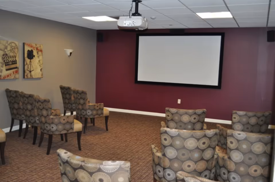 A small theater room with patterned armchairs arranged in rows facing a large blank projection screen mounted on a maroon wall. The room has a beige carpet, gray walls with two framed movie-themed artworks, and a ceiling-mounted projector.