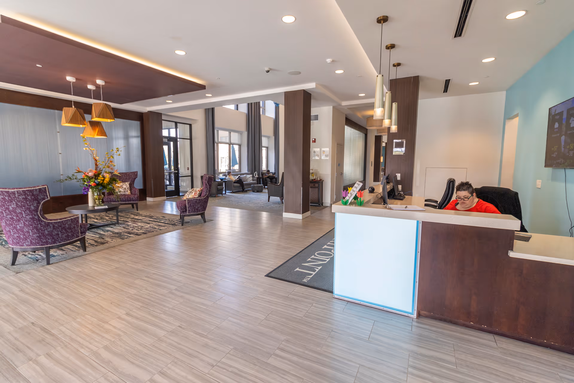 A spacious and modern reception area in a senior living facility with a front desk on the right where a staff member is seated. The room features light wood flooring, purple upholstered chairs around a coffee table with a floral arrangement, and large windows letting in natural light. Pendant lights hang from the ceiling, and a television is mounted on the wall behind the desk.