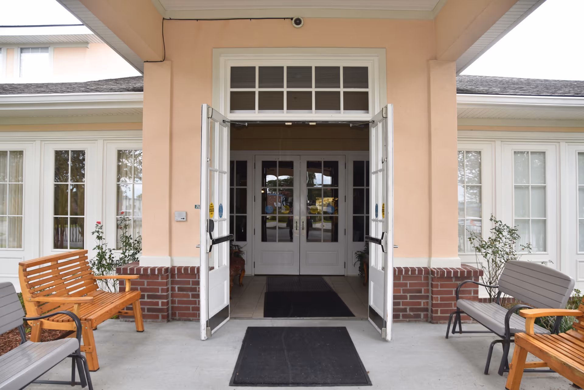Front entrance of a senior living facility with open double doors, benches, potted plants, and large windows.