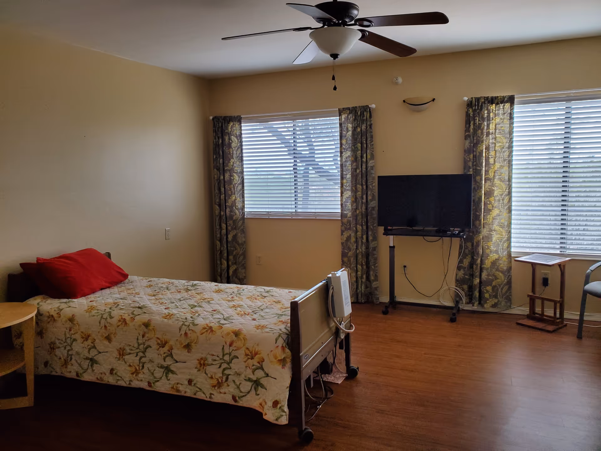 A simple bedroom in a senior living facility with a single bed covered in a floral quilt and a red pillow. The room has two windows with floral curtains, a ceiling fan, a TV mounted on a stand, a small side table, and a chair. The floor is wooden, and the walls are painted beige.