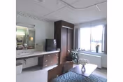 A nursing facility room with a bed, a wooden wardrobe, a TV on a dresser, a sink with a mirror above it, a small table with a flower vase, and a large window letting in natural light.