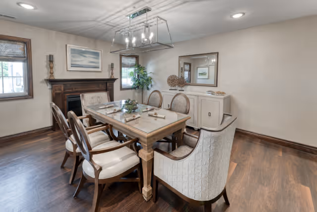 A dining room with a rectangular wooden table surrounded by six upholstered chairs. The table is set with napkins and decorative items. The room features hardwood floors, two windows with blinds, a wooden fireplace mantel, a sideboard with a mirror above it, and a modern chandelier hanging from the ceiling.