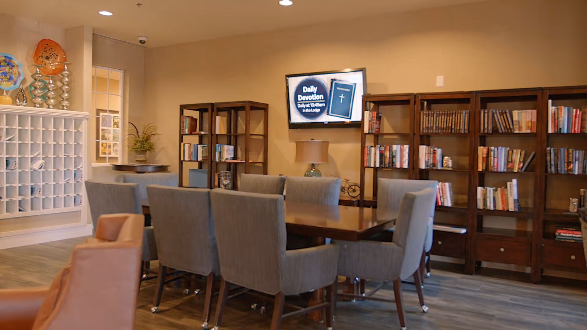 A cozy interior room with a large wooden table surrounded by eight gray upholstered chairs. Behind the table are several dark wooden bookshelves filled with books. A flat-screen TV mounted on the wall displays a notice for a daily devotion at 10:45 am in the lodge. Decorative items and a lamp are also visible on the shelves and table. The room has warm lighting and a wooden floor.