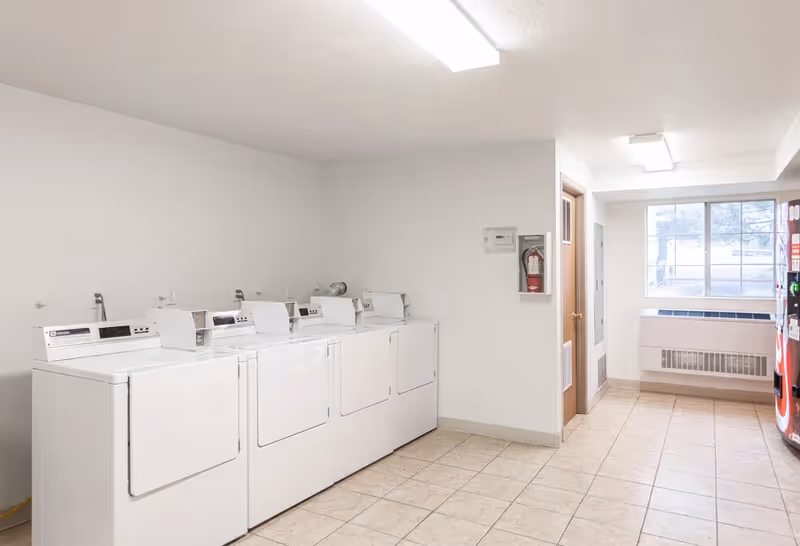 A laundry room with four white washing machines and dryers lined up against a white wall. The room has tiled flooring, a fire extinguisher mounted on the wall, a wooden door, a large window letting in natural light, and a vending machine on the right side.