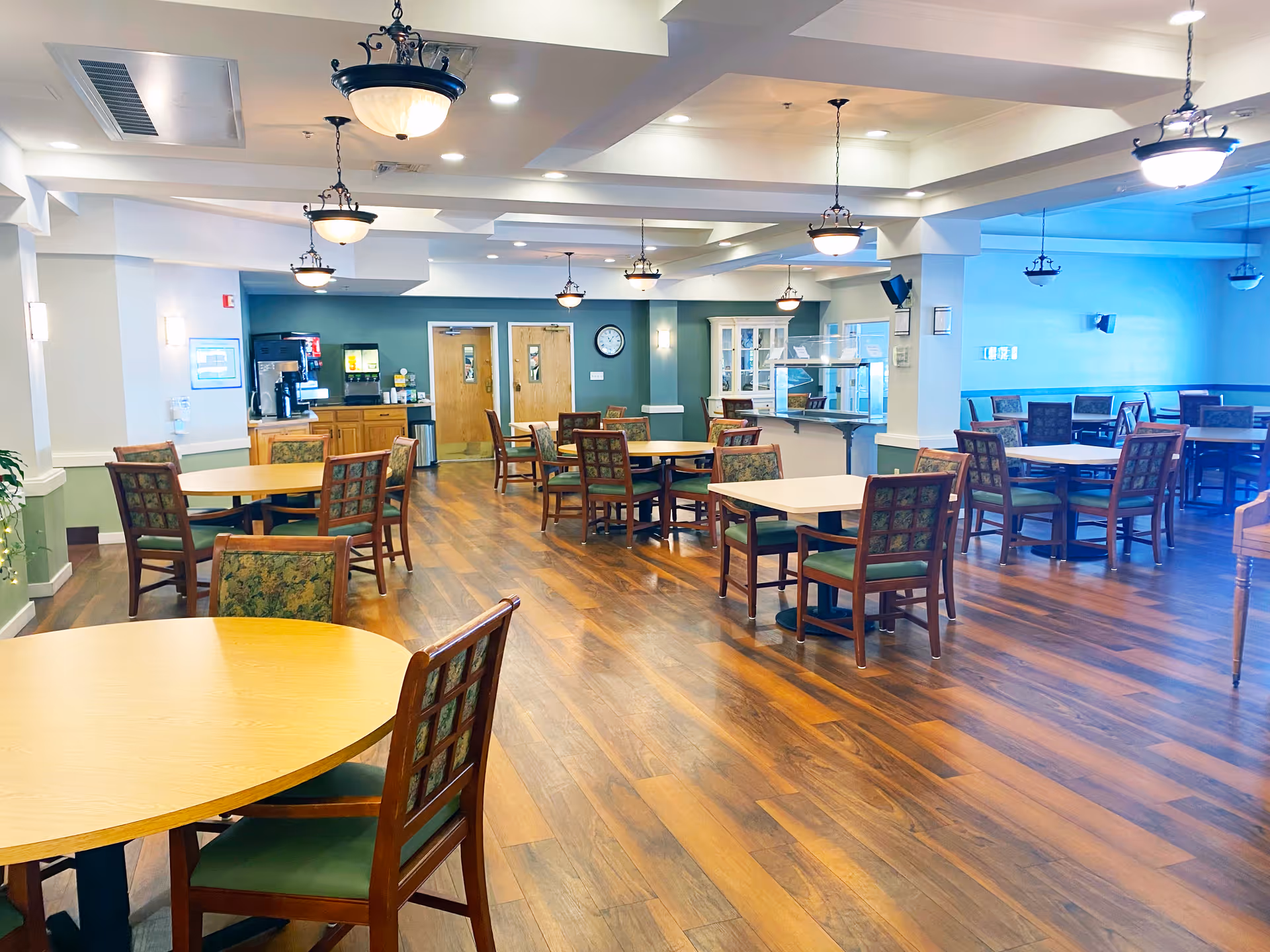 A spacious dining area in a senior living facility with multiple round and square wooden tables surrounded by chairs with green cushions and floral-patterned backs. The room features wooden flooring, hanging light fixtures, a beverage station with coffee and juice dispensers, and a clock on the wall. The walls are painted in soft green and white tones.