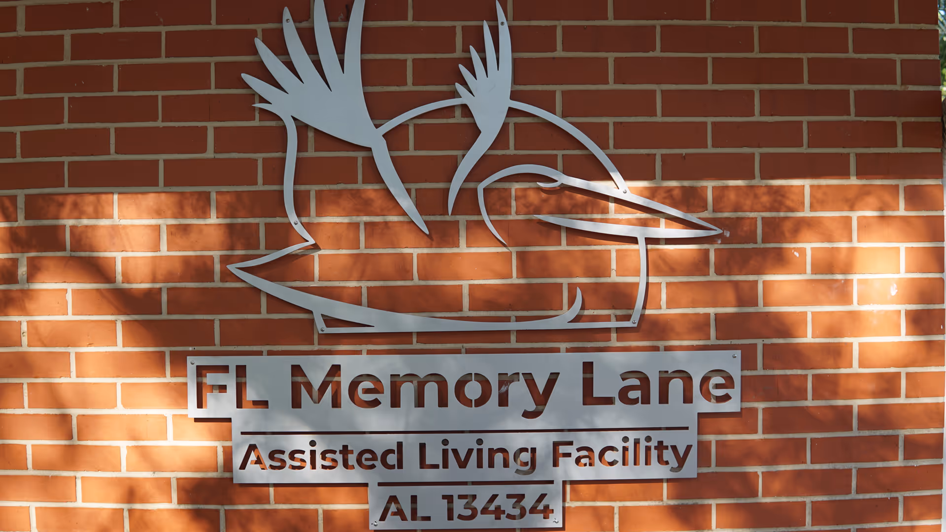 Metal sign mounted on a red brick wall displaying a stylized bird logo above the text 'FL Memory Lane Assisted Living Facility AL 13434'.