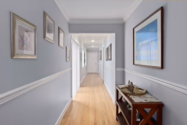 A long, narrow hallway with light gray walls and white trim. The floor is light wood, and framed pictures hang on both walls. At the end of the hallway, there is a white door. On the right side near the foreground, there is a wooden console table with decorative items on top.