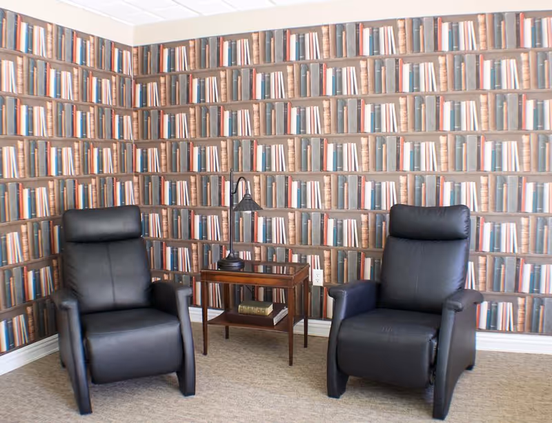 Two black leather recliners flank a wooden side table with a lamp against wallpaper printed to look like bookshelves.