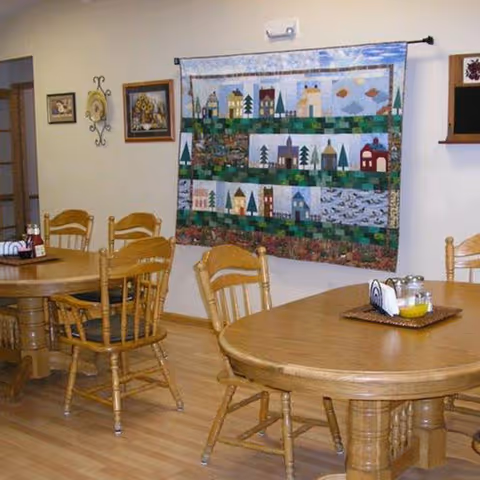 Wooden dining tables and chairs in a communal dining room with a colorful quilt hanging on the wall.