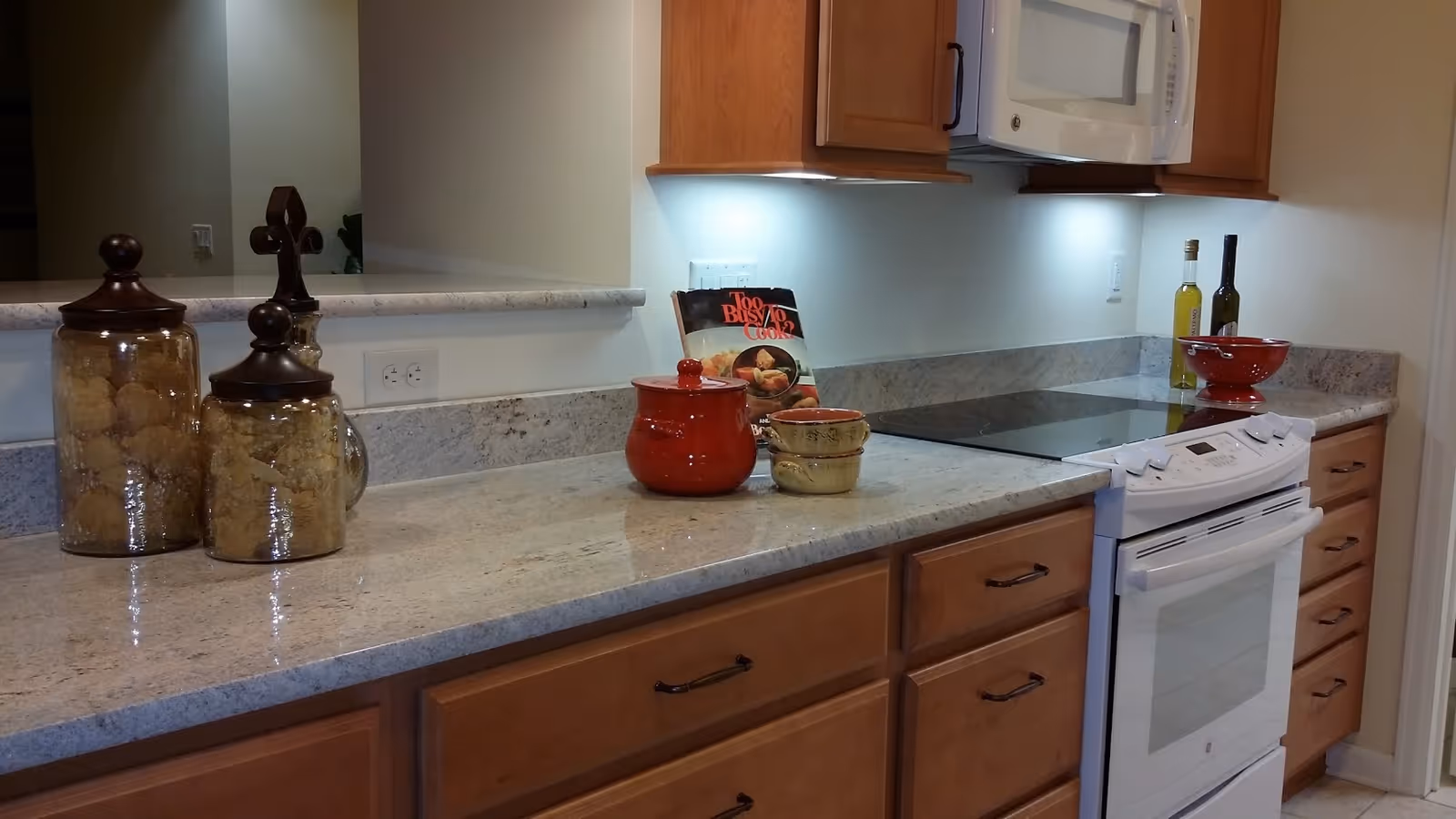 A modern kitchen countertop with wooden cabinets, a white electric stove and oven, a white microwave mounted above the stove, and various kitchen items including glass jars with cookies, a red pot, a cookbook, two bottles of oil, and a red bowl.