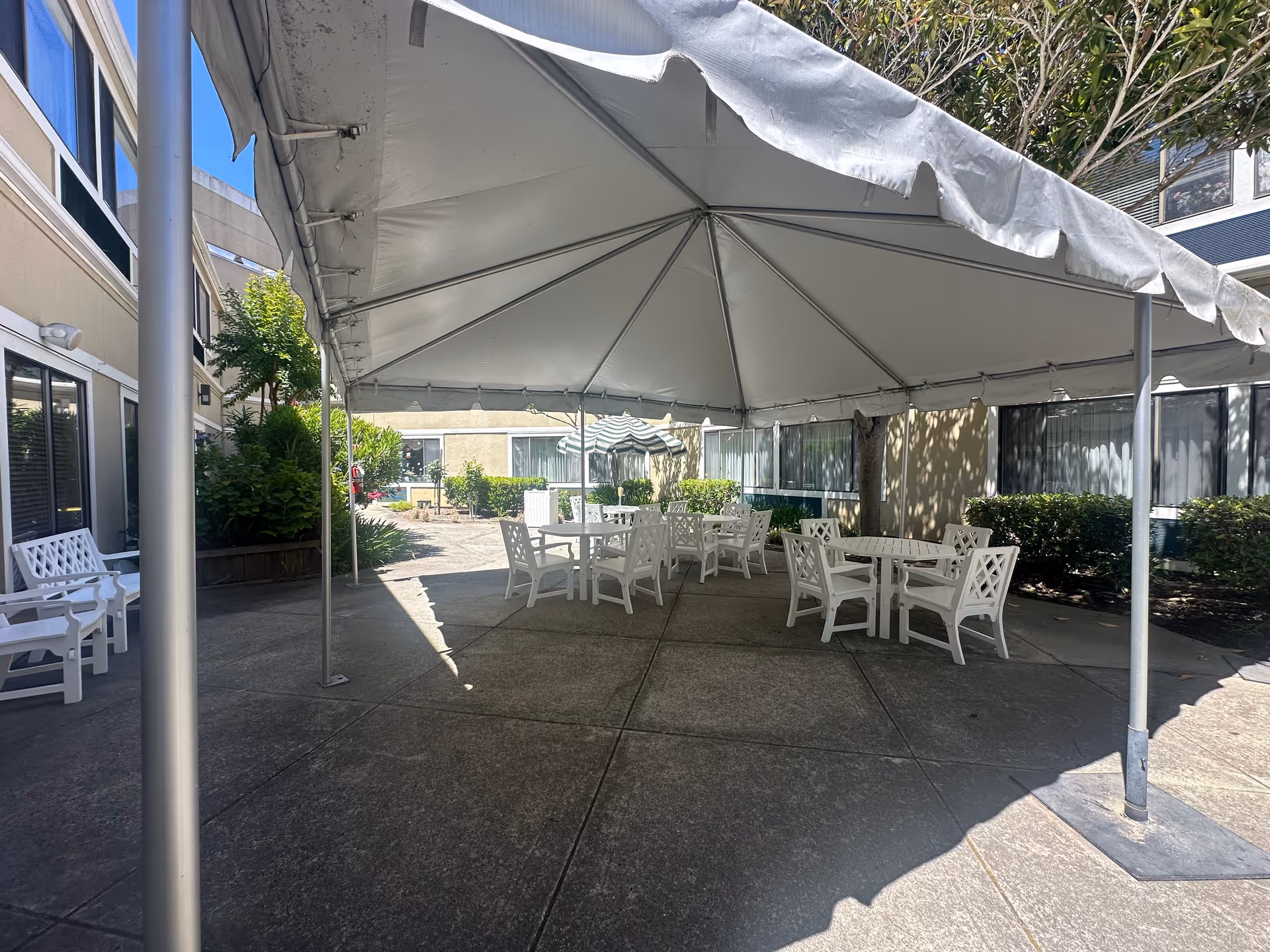 Outdoor patio area at Rossmoor Post Acute with a large white canopy tent covering several white tables and chairs. The patio is surrounded by bushes and windows of the building, with additional seating and greenery visible in the background.