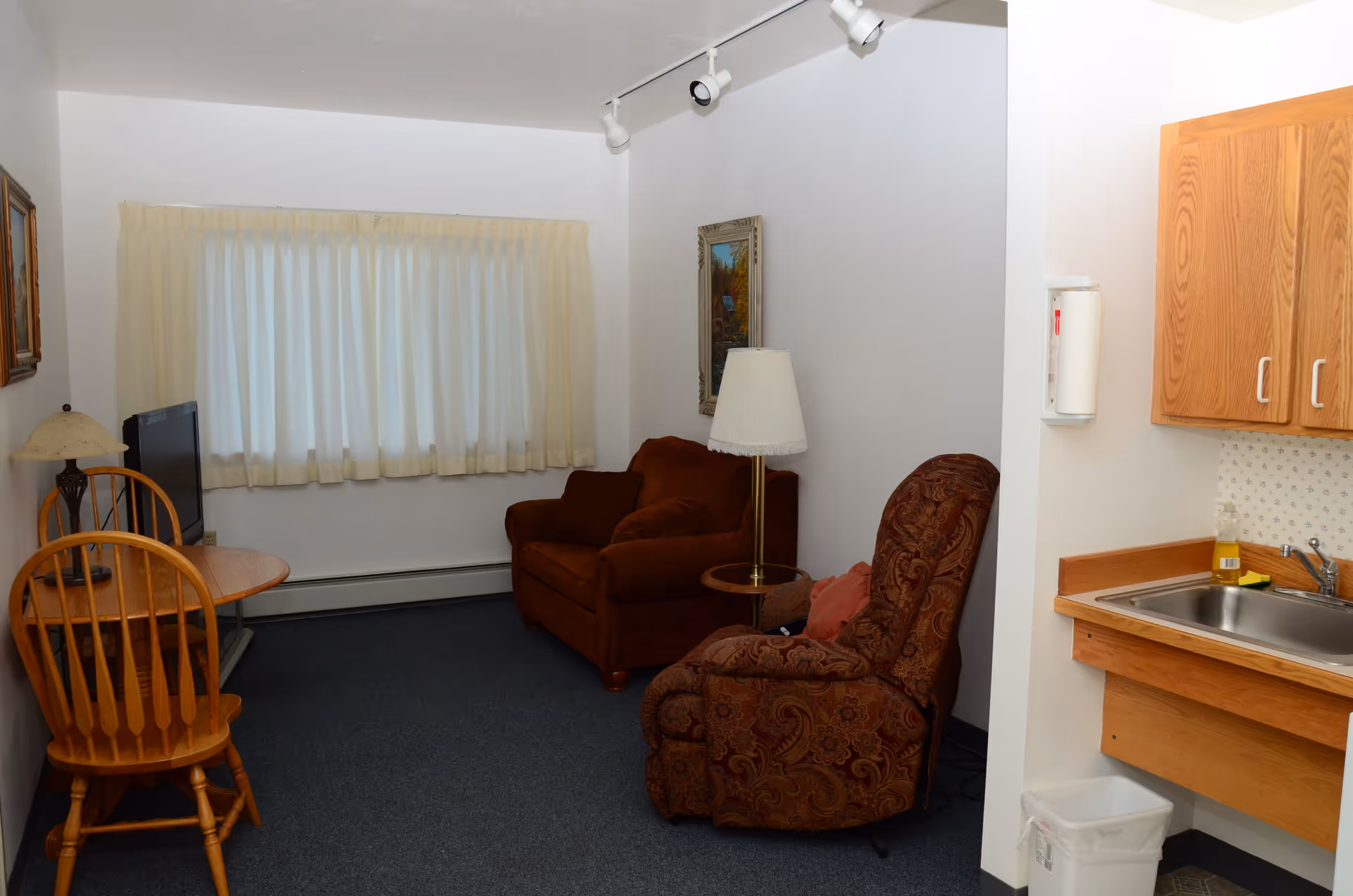 A small living area in a senior living facility with a brown upholstered armchair, a patterned recliner chair, a floor lamp, a small wooden table with two wooden chairs, a TV on a stand, and a kitchenette with a sink and wooden cabinets. The room has white walls, a window with cream-colored curtains, and blue carpeted flooring.
