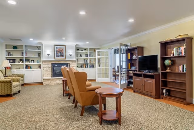 A spacious living room area with a row of four brown armchairs facing a stone fireplace with a framed picture above it. The fireplace is flanked by built-in white bookshelves filled with books and decorative items. To the right, there is a wooden entertainment center with a flat-screen TV and additional bookshelves. The room has beige walls, recessed ceiling lights, and a carpeted floor. A doorway with glass French doors leads to another room in the background.