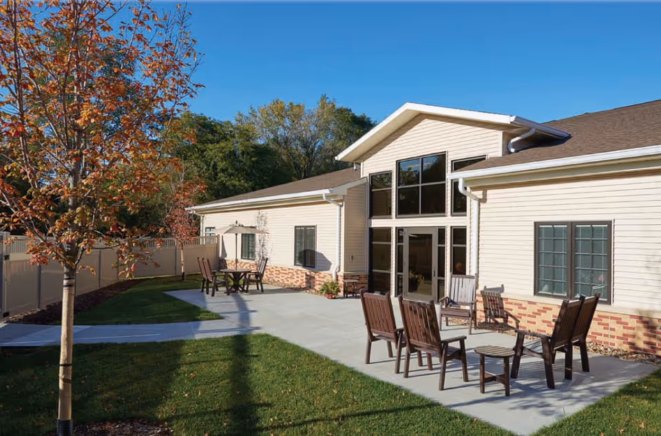 Outdoor patio area of a senior living facility with several wooden chairs and small tables arranged on a concrete surface. The building has beige siding with brick accents and large windows. There is a tree with autumn-colored leaves and a white fence enclosing the area, under a clear blue sky.
