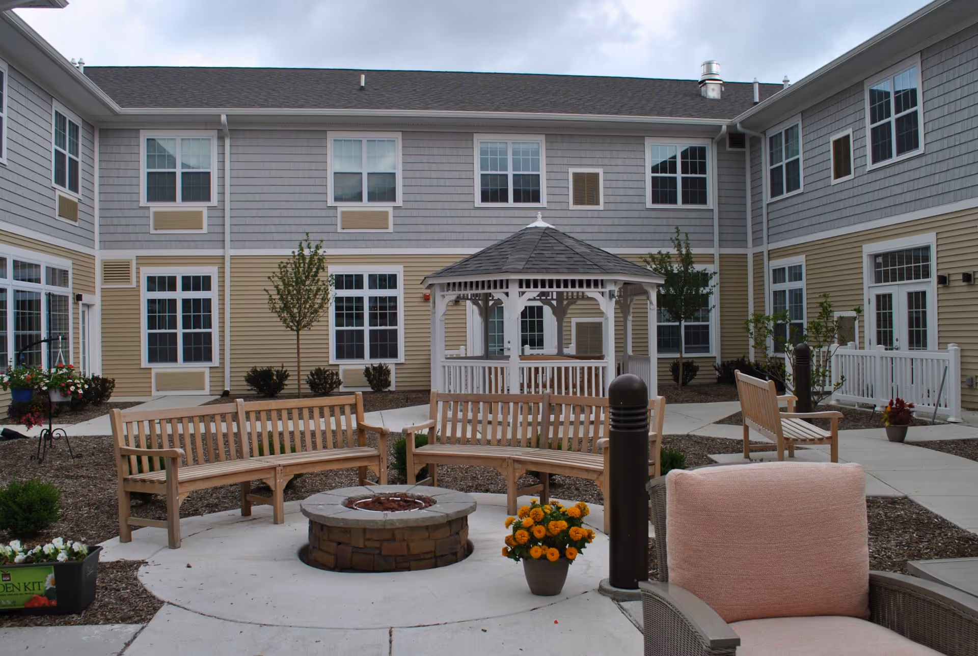 Courtyard with wooden benches around a central fire pit, a white gazebo, potted flowers, and a two-story building surrounding the area.