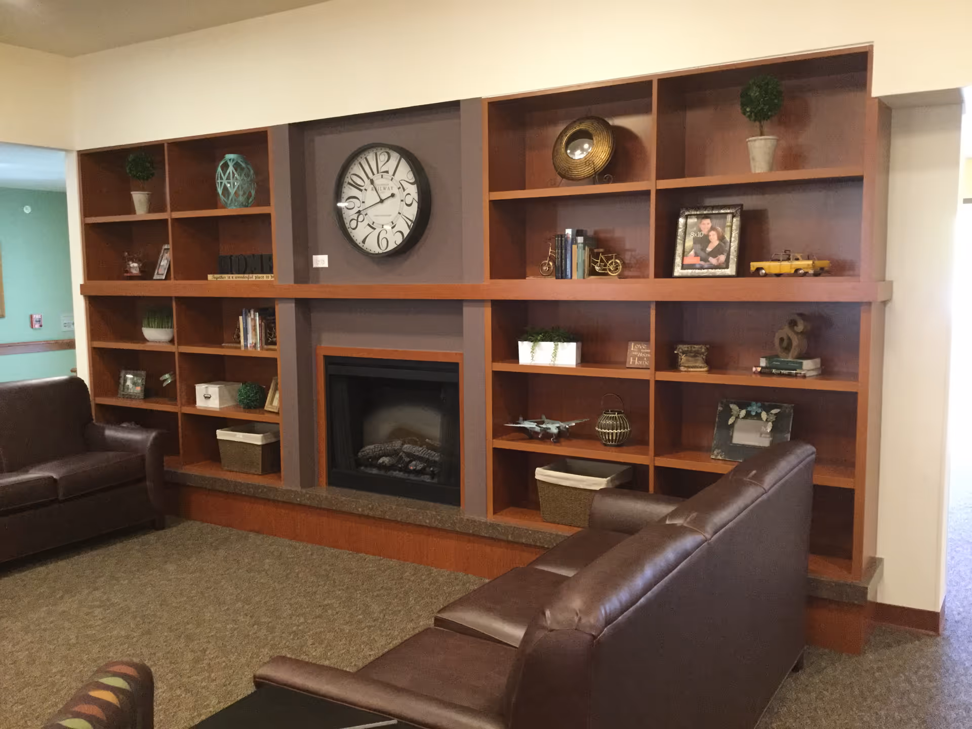 A community living room with wooden built-in shelves and a fireplace, leather sofas, a clock, and decorative items.