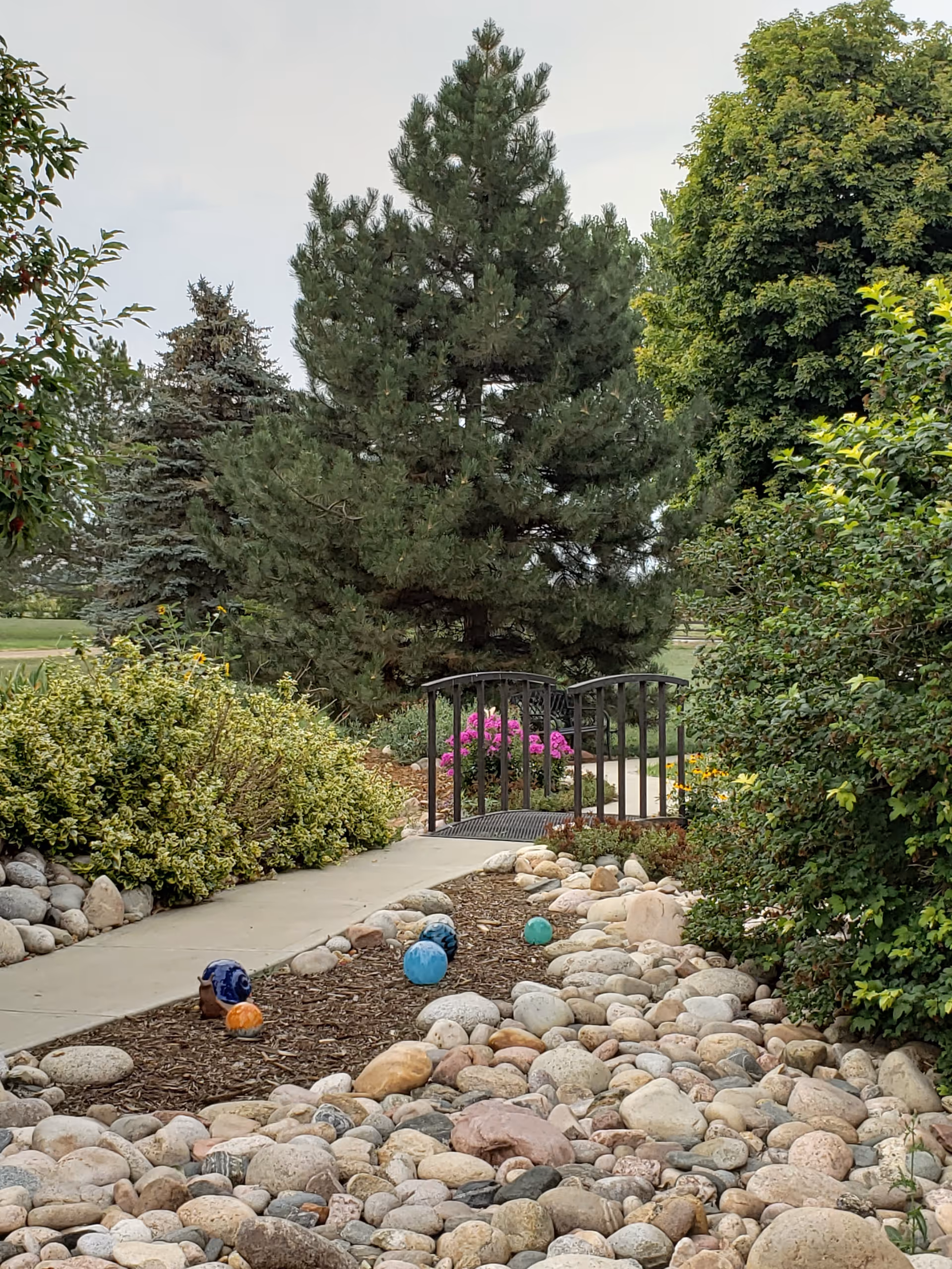 A landscaped outdoor path leading to a small decorative bridge surrounded by rocks, shrubs, flowering plants, and large trees.