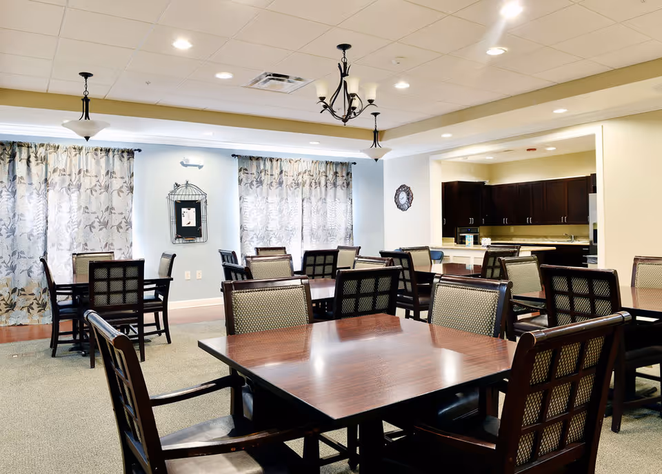 A well-lit dining room with multiple wooden tables and cushioned chairs arranged neatly. The room features large windows with floral-patterned curtains, ceiling lights, and a chandelier. In the background, there is an open kitchen area with dark wooden cabinets and a countertop.