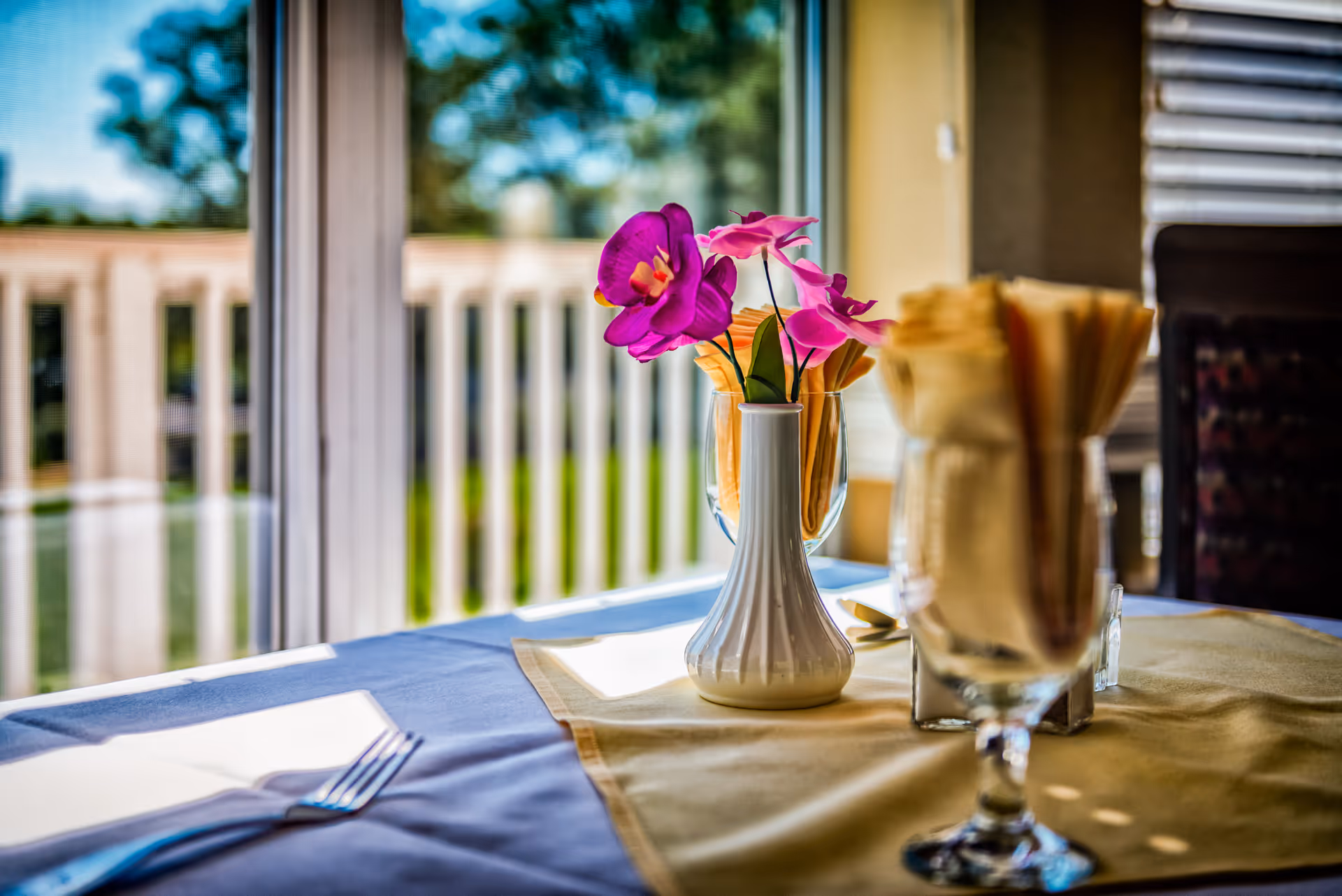 A sunlit dining table by a window with a small white vase holding purple flowers and glasses with folded napkins.