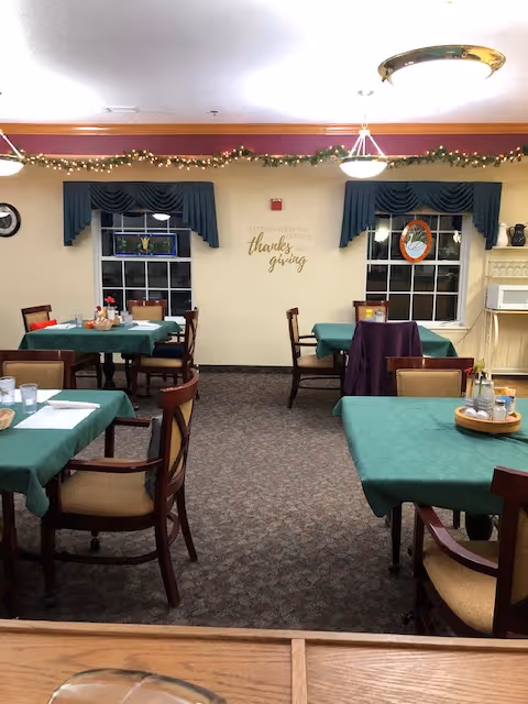 Dining room with several tables covered in green tablecloths and set with napkins, cups, and condiments. The room has carpeted floors, two windows with blue valances, and a wall decoration that says 'Give thanks with a grateful heart Thanksgiving'. There are string lights hanging along the top of the wall and a ceiling light fixture.