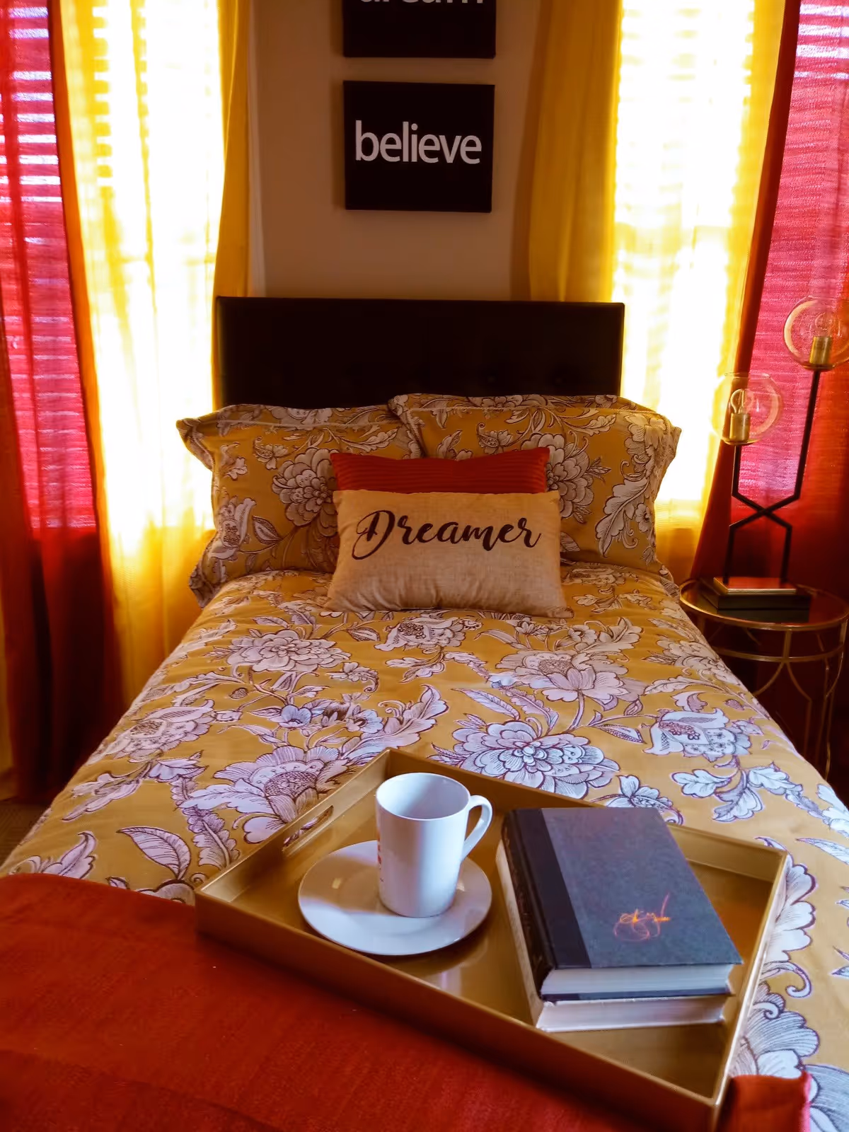 A bright bedroom with a yellow floral bedspread, decorative pillows including one that says 'Dreamer', a tray holding a cup and books, and yellow and red curtains.