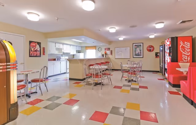 A retro-style dining area with a kitchen in the background. The room features a colorful tiled floor with red, yellow, gray, and white squares. There are several metal tables and chairs with red cushions, a red booth seating area, a Coca-Cola vending machine, and a jukebox. The walls are decorated with vintage Coca-Cola posters and a whiteboard.