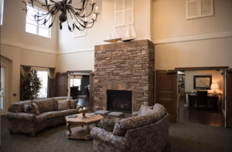 A cozy senior living facility common area with two patterned sofas arranged around a wooden coffee table in front of a large stone fireplace. The room has high ceilings with a decorative chandelier, large windows, and doorways leading to other rooms.
