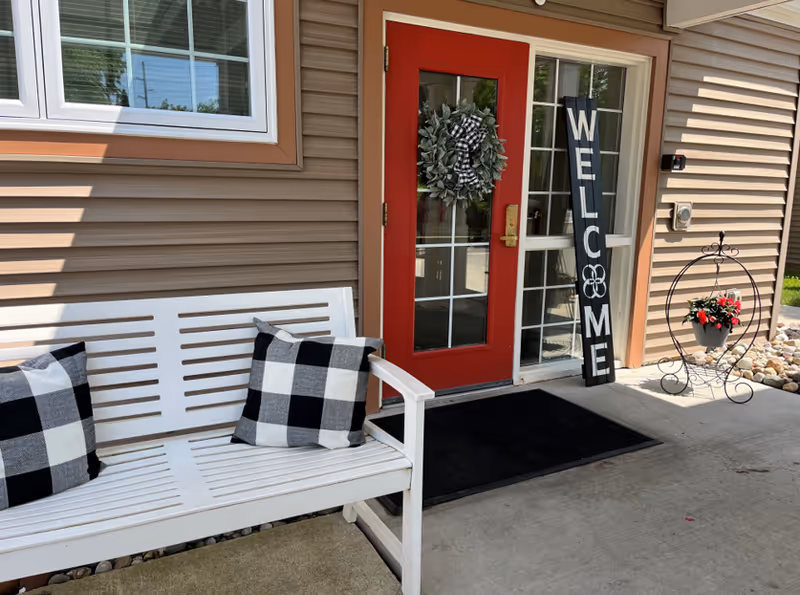 Entrance to a facility with a red door decorated with a wreath and a tall black welcome sign leaning against the wall. A white bench with two black and white checkered pillows is placed beside the door. There is a small decorative plant stand with a pot of red flowers on the right side of the entrance.