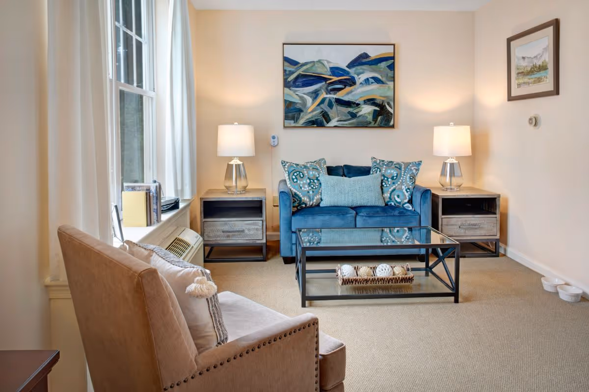 A cozy living room with a blue loveseat adorned with patterned and solid pillows, two matching side tables each with a lamp, a glass coffee table with decorative items, a beige armchair with a pillow, and two framed paintings on the walls. There is a window with white curtains and an air conditioning unit below it.