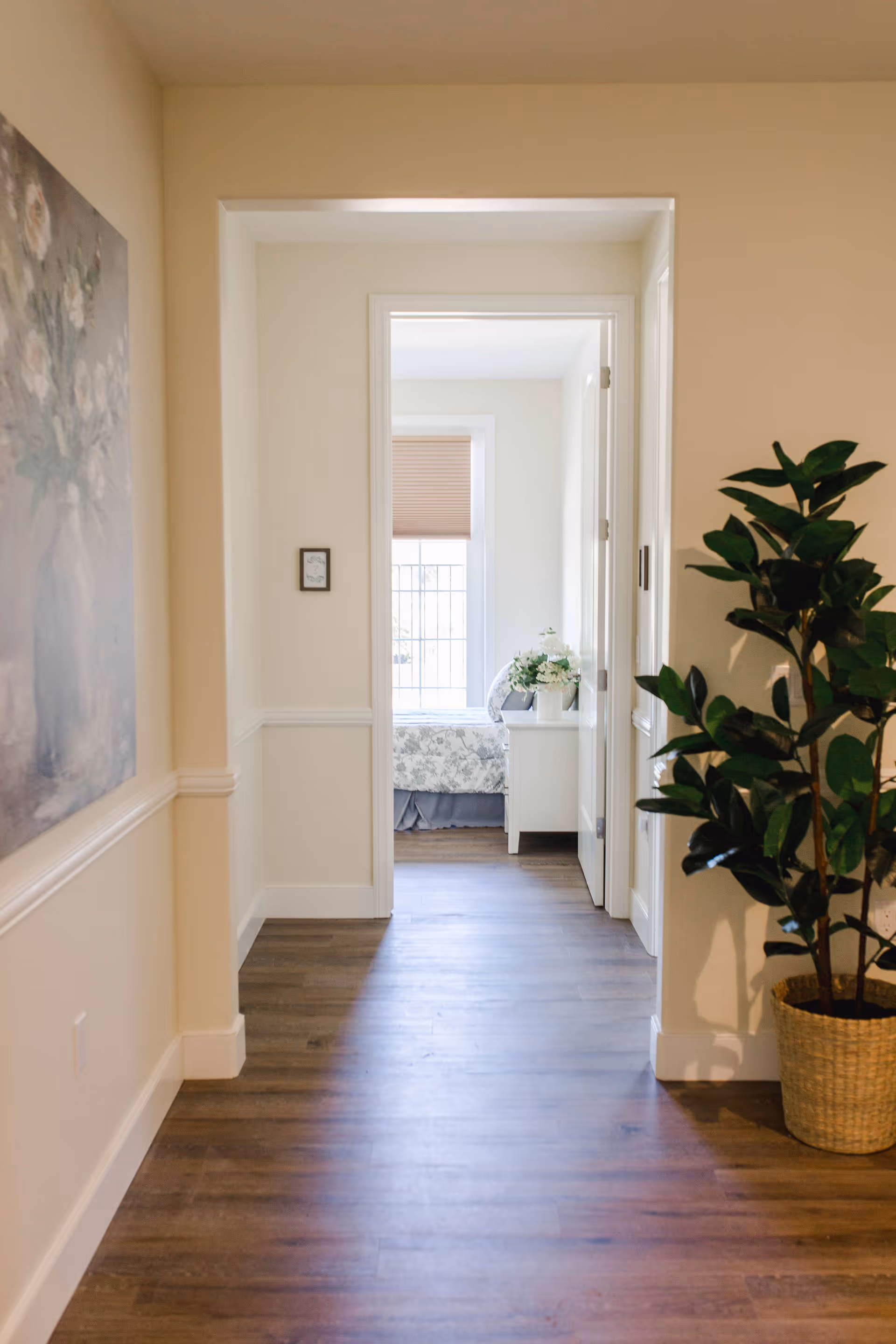 View down a hallway with wooden flooring leading to a bedroom. The bedroom has a window with a blind, a bed with floral bedding, and a white nightstand with a flower arrangement. A large potted plant is on the right side of the hallway, and a floral painting hangs on the left wall.