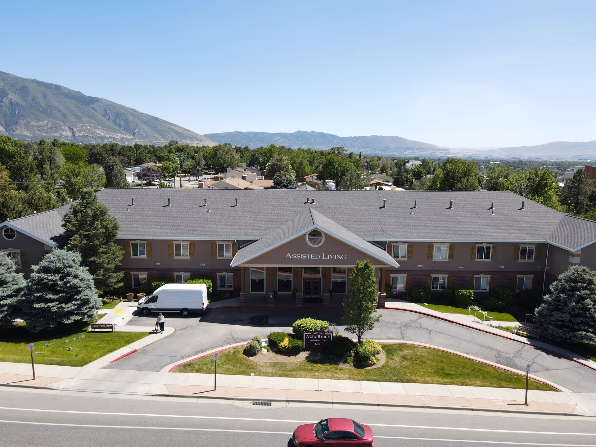 Front exterior view of Alta Ridge Assisted Living of Sandy, a two-story building with a gray roof and brown walls, surrounded by trees and greenery. A white van is parked near the entrance, and a person is walking on the driveway. Mountains and a clear blue sky are visible in the background.