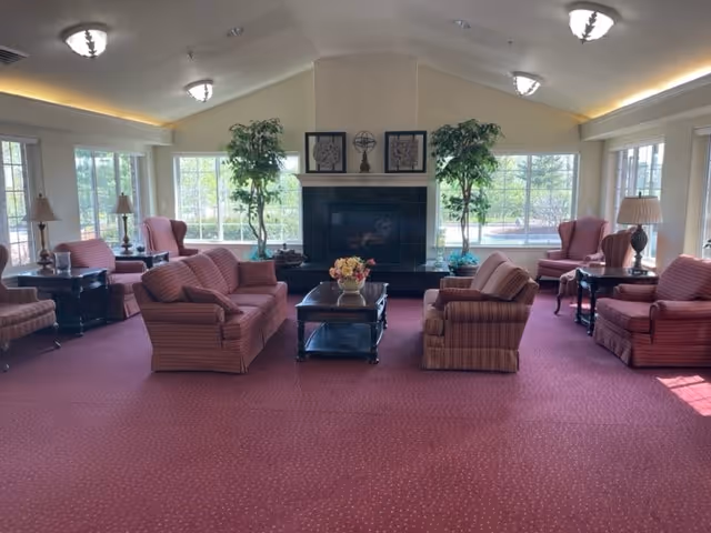 Spacious senior living lounge with upholstered sofas and armchairs arranged around a central coffee table and fireplace, flanked by large windows and potted plants.