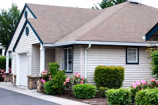Exterior view of a single-story building with beige siding and a brown shingled roof. The building has a garage door, a window, and is surrounded by green shrubs and pink flowers. Trees are visible in the background.