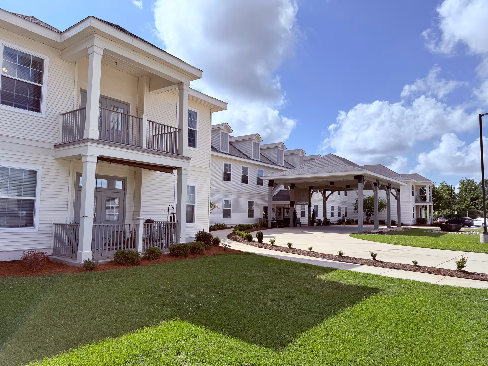 Exterior view of a senior living facility with white siding, multiple windows, balconies, and a covered entrance area. The building is surrounded by a well-maintained lawn and landscaping under a partly cloudy blue sky.