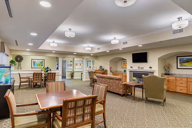 A spacious senior living facility common area with carpeted floors and neutral-colored walls. The room features multiple seating arrangements including a wooden table with four chairs in the foreground, a floral-patterned sofa, and armchairs near a fireplace with a mounted flat-screen TV above it. There are cabinets along the walls, decorative artwork, a fish tank on the left, and ceiling lights providing bright illumination.