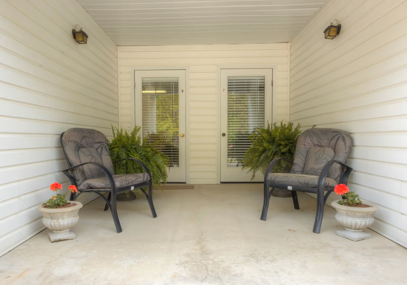 A covered porch area with two cushioned chairs facing each other, each accompanied by a potted red flower and a large green fern behind. Two white doors with glass panels and blinds are visible in the background, numbered 9 and 7. The walls and ceiling are white with horizontal paneling, and there are two wall-mounted lights above the chairs.